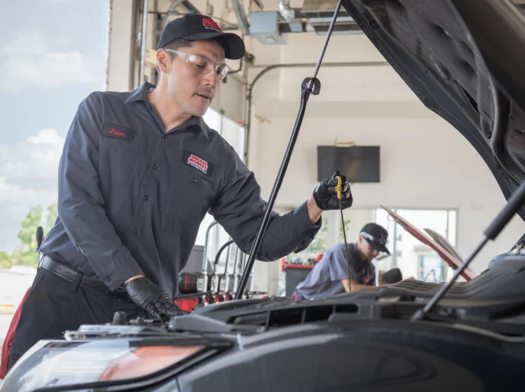 A Grease Monkey mechanic in uniform inspects a car engine with the hood open inside an auto repair shop, possibly preparing for Fluid Exchange Services; another worker is in the background.