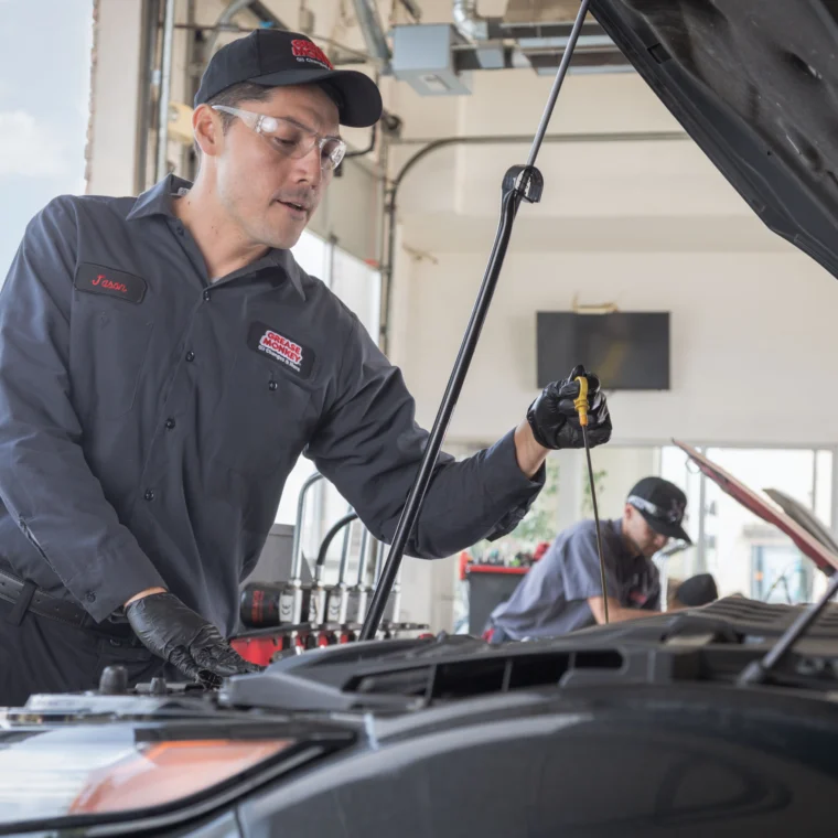A Grease Monkey mechanic in uniform inspects a car engine with the hood open inside an auto repair shop, possibly preparing for Fluid Exchange Services; another worker is in the background.