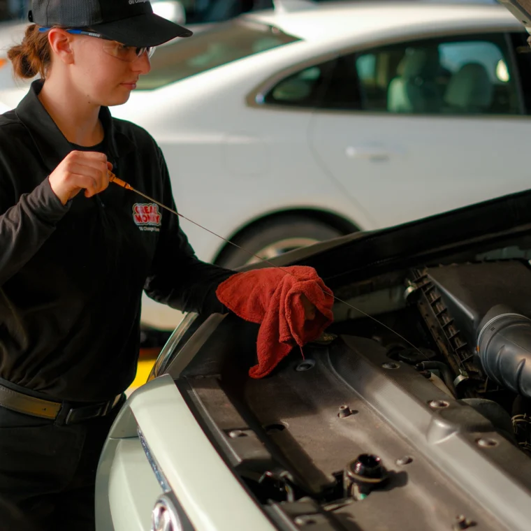 An auto mechanic in uniform checks the oil level of a car engine with a dipstick while holding a red cloth, ensuring top-notch Fluid Exchange Services.