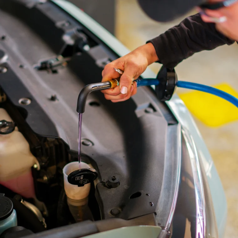 A person pours coolant into a car's windshield washer fluid’s reservoir using a hose, with the hood open in a garage setting—a common step in routine Fluid Exchange Services.