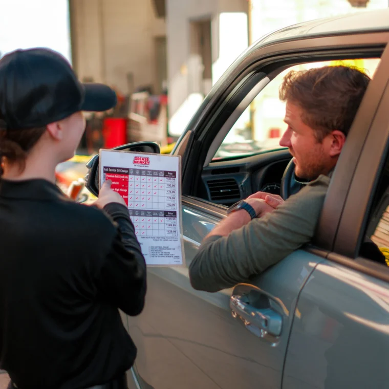 A person in uniform shows a checklist to a man in his car at a drive-through service location specializing in Fluid Exchange Services.