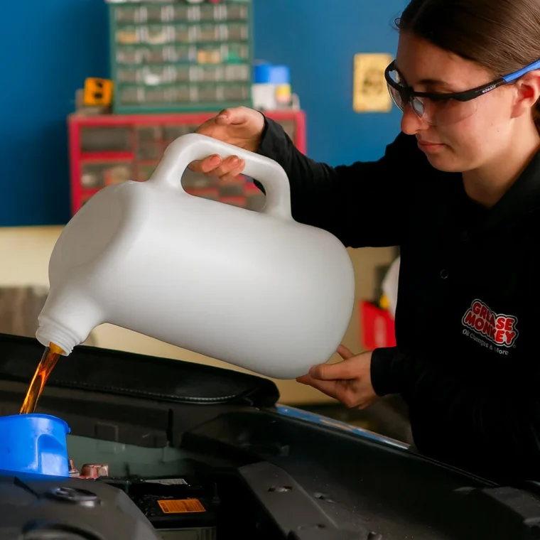 A person wearing safety glasses pours oil from a large container into a car engine using a blue funnel.
