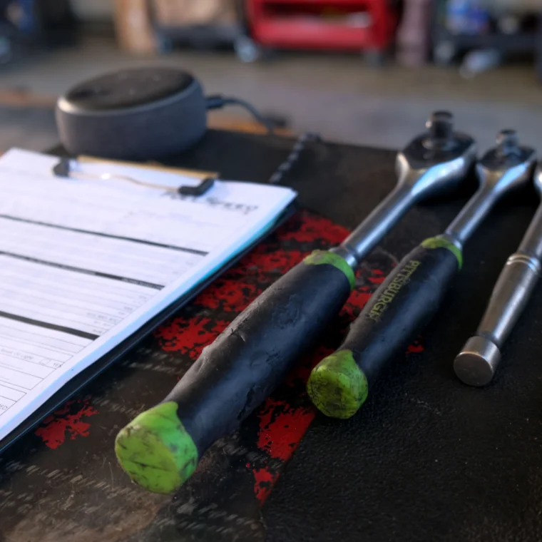 A clipboard with paperwork sits next to various ratchets and sockets on a workbench in a garage setting.