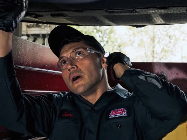 A mechanic wearing safety glasses and gloves examines the underside of a vehicle in a workshop, with tools and equipment visible in the background—a scene that reminds us to thank a mechanic for keeping our vehicles safe and reliable.
