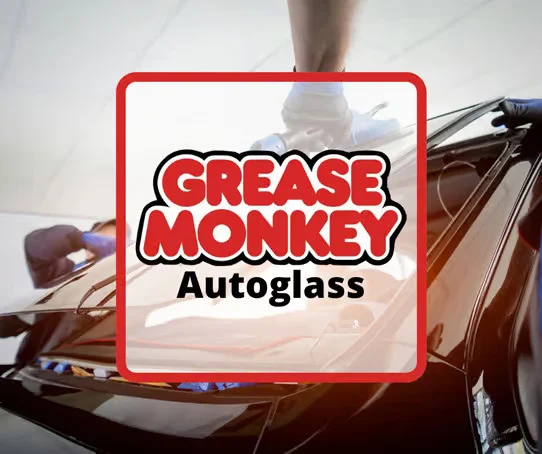 Technicians perform Auto Glass Repair as they install a windshield on a black car in Idaho, with a "Grease Monkey Autoglass" logo overlayed in the center.