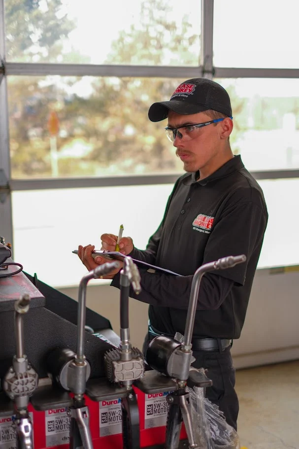 A mechanic in uniform and glasses writes on a clipboard in a garage, with automotive tools and engine parts visible in the foreground.