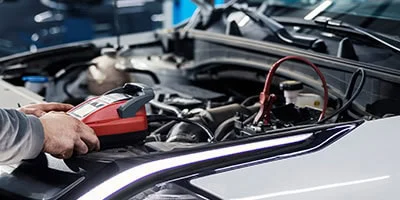 A Grease Monkey tests a car battery under the hood with diagnostic equipment and jumper cables, ensuring the battery is checked for optimal performance.