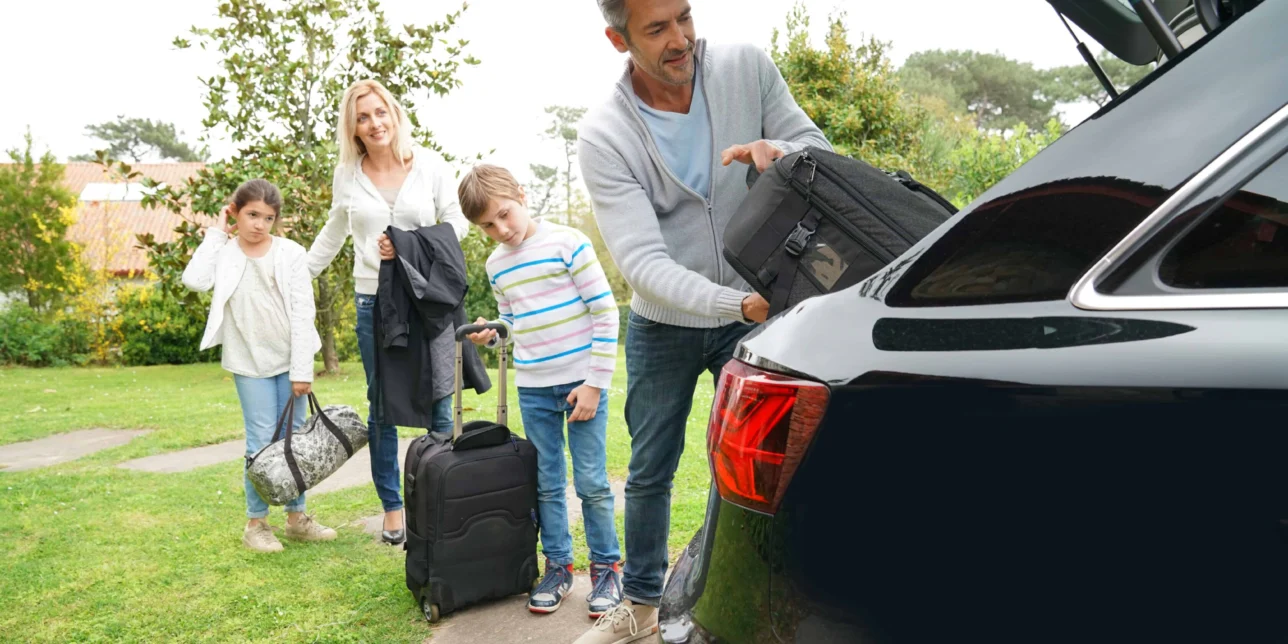 A man loads a suitcase into a car trunk while a woman and two children with luggage stand nearby on a grassy area, busy with car preparation for their road trip.