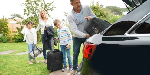 A man loads a suitcase into a car trunk while a woman and two children with luggage stand nearby on a grassy area, busy with car preparation for their road trip.