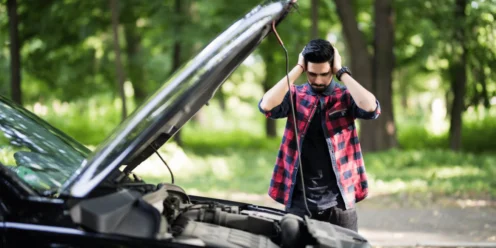 A man stands in front of a car with the hood open, holding his head in frustration, realizing the importance of regular car maintenance for keeping things running smoothly, with trees in the background.