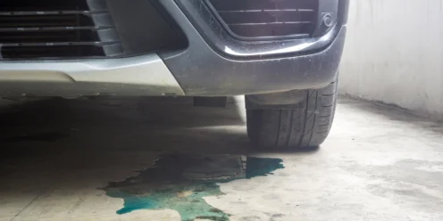 A close-up of a car's front bumper and tire shows a greenish fluid leaking onto the concrete floor—one of the common car leaks that highlights the need for regular car maintenance.