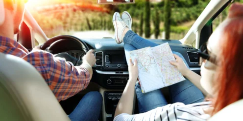 Two people in a car driving; the driver holds the wheel while the passenger, with feet up on the dashboard, looks at a map. Sunlight is shining through the windshield—a reminder of the importance of summer car care and how to protect your car from heat.