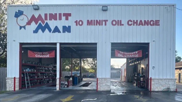 A white auto service building with "MINIT MAN 10 MINIT OIL CHANGE" on the front, two open garage bays, and shelves with supplies visible inside.
