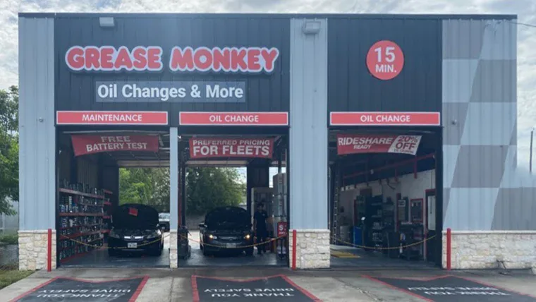 Exterior of a Grease Monkey auto service center with three service bays, two cars inside, and signage for oil changes, battery tests, fleet services, and rideshare readiness.