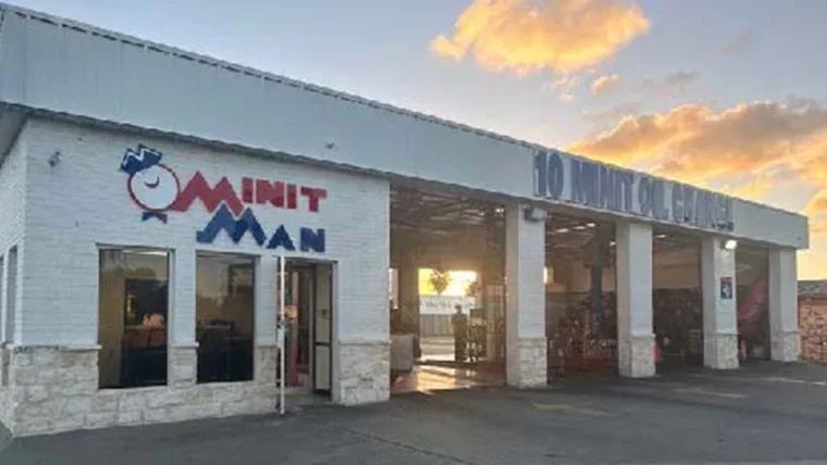Exterior view of an auto service shop with "10 MINIT OIL CHANGE" and "MINIT MAN" signs on the building, with sunset skies in the background.