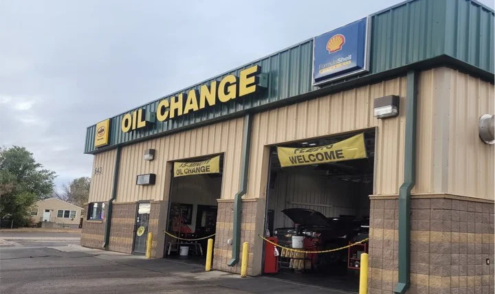 A tan and green automotive shop with large "OIL CHANGE" sign and two open service bays, one with a car inside and a yellow "WELCOME" banner above.