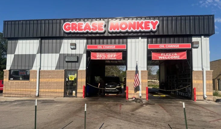A Grease Monkey auto service shop with two open service bays, a car inside, red signage, and an American flag outside. A “25% off” banner hangs over one bay.