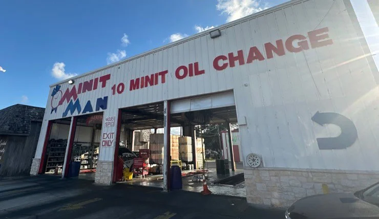 Exterior of an auto service shop with “10 MINIT OIL CHANGE” painted above open garage doors; cars and equipment visible inside.
