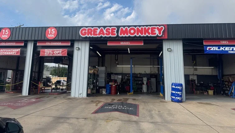 Exterior view of a Grease Monkey auto service center with open service bays, signage for oil changes, tires, brakes, and a 25% off discount sign displayed.
