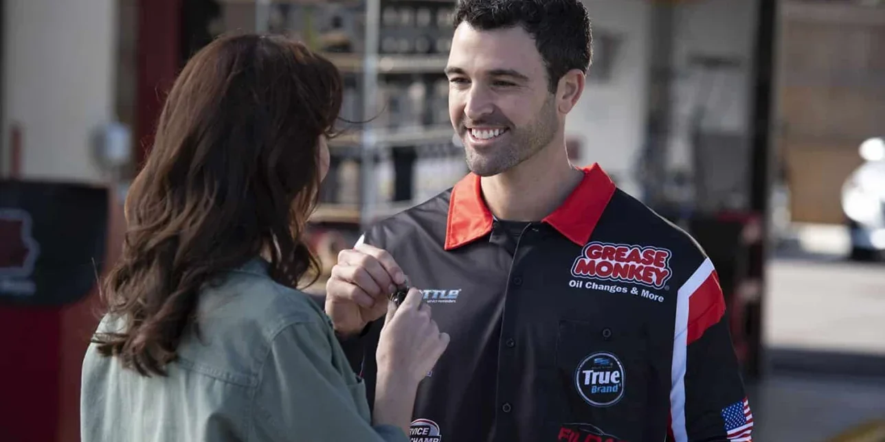 A mechanic in a Grease Monkey uniform hands a set of keys to a woman at an auto service center, echoing the spirit of Westward Expansion as they prepare for new journeys on the open road.