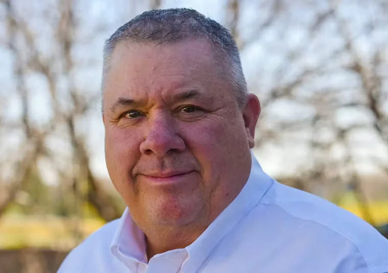 A middle-aged man with short gray hair wearing a white collared shirt, standing outdoors with leafless trees in the background.