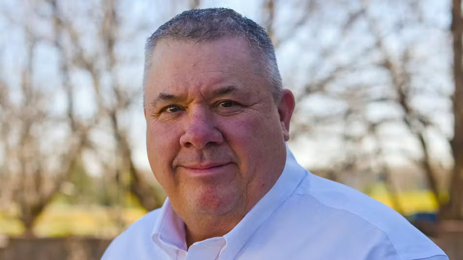 A middle-aged man with short gray hair wearing a white collared shirt, standing outdoors with leafless trees in the background.