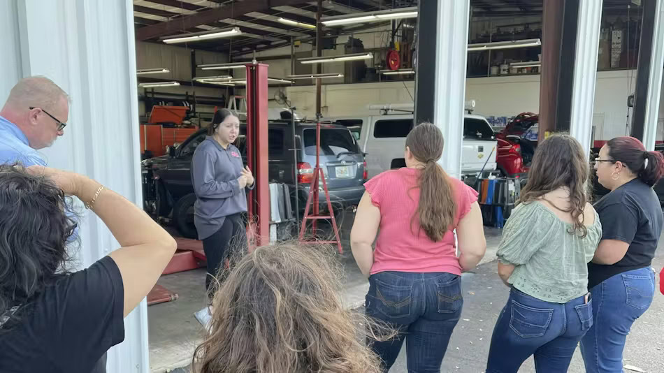 A group of people stand in an auto repair shop listening to a woman speaking near a car lifted on a hydraulic lift.