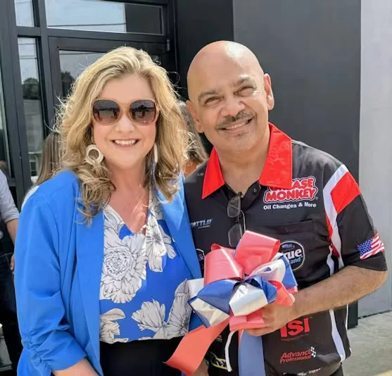 Two people pose together outside; the woman wears sunglasses and a blue blazer, while the man holds a red, white, and blue ribbon and wears a black sponsor-branded shirt.