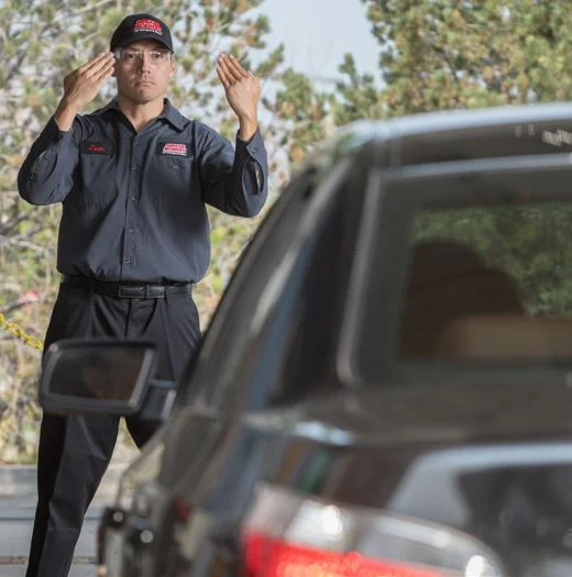 A uniformed man stands in front of a car, directing the driver with hand signals in an outdoor setting.