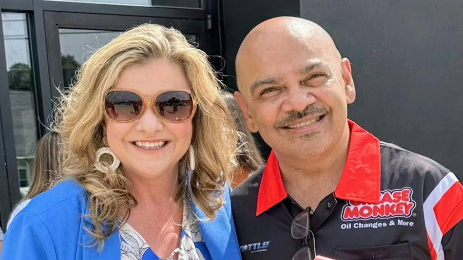 Two adults standing side by side, smiling at the camera. The woman is wearing sunglasses and a blue blazer; the man is wearing a “Grease Monkey” shirt.