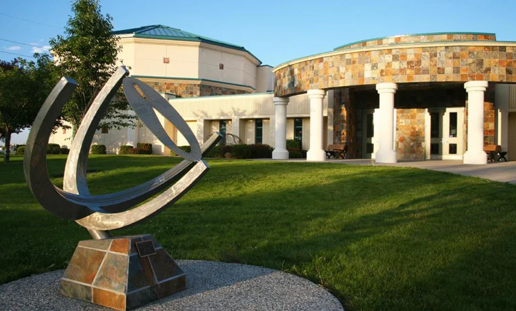 A modern metal sundial sculpture stands on a stone base in front of a building with white columns and stone accents near Grease Monkey Oil Change in Twin Falls, Idaho, highlighting the importance of engine protection.