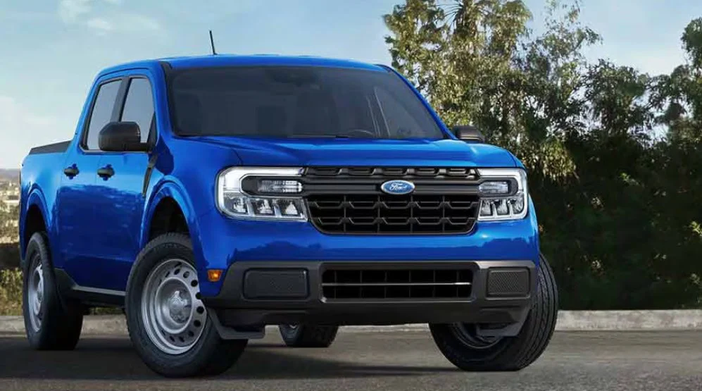 A blue Ford pickup truck is parked outdoors on a paved surface at Grease Monkey in Jerome, Idaho, with green trees and shrubbery in the background—ready for engine long life maintenance.
