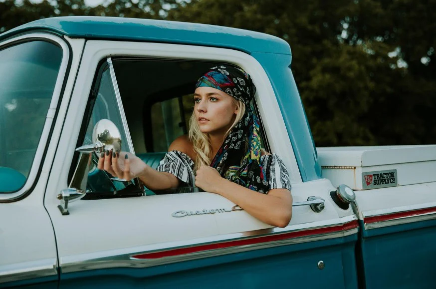 A woman wearing a colorful headscarf sits in the driver’s seat of a vintage blue and white pickup truck in Idaho Falls, adjusting the side mirror as she prepares for her next trip and engine oil care check.