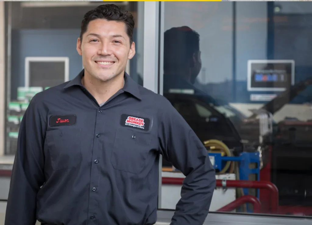 A smiling man in a dark uniform shirt stands in front of a window at an auto repair shop in Rexburg, ready to help with oil changes and car maintenance.