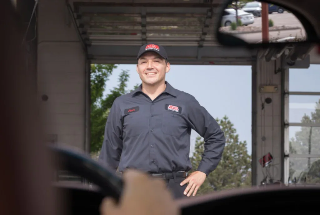 A mechanic in a black uniform and cap stands smiling in front of an open garage door in Pocatello, Idaho, ready to help save your engine with a quick oil change—viewed from inside a vehicle.