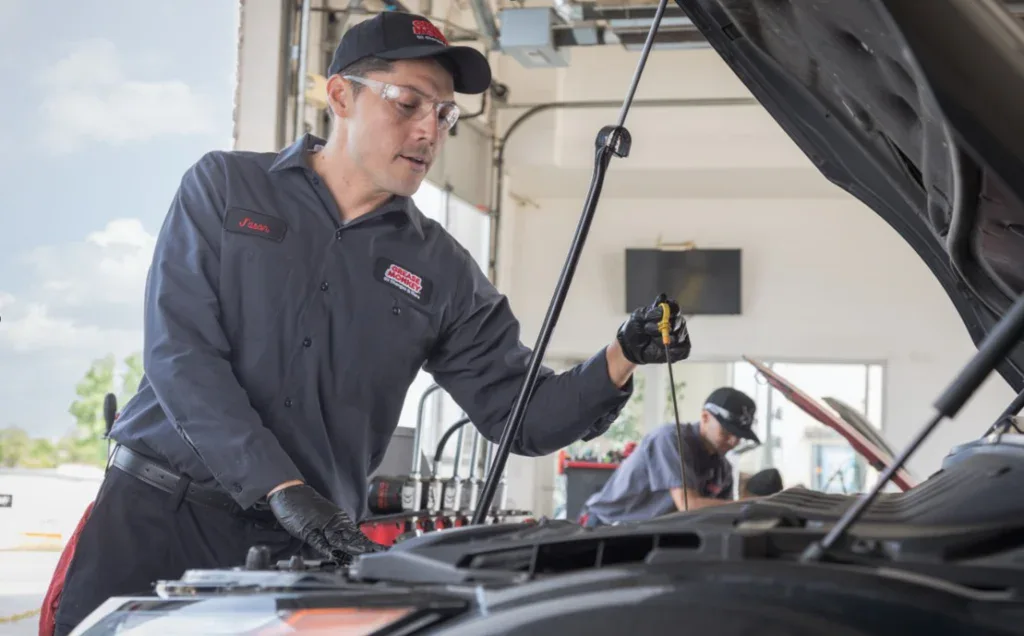 A Grease Monkey auto mechanic in uniform and gloves checks the oil level under the hood of a car in Grease Monkey 2100 Channing Way, Idaho Falls, Idaho service garage in Idaho Falls, with another worker visible in the background, ensuring engine longevity by using an oil change near me and using synthetic blend oil for engine longevity in Idaho Falls, Idaho.