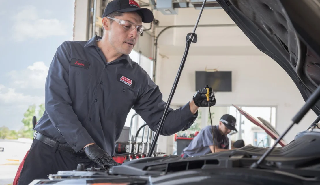 An auto mechanic in uniform and gloves checks the oil level under the hood of a car in a service garage in Idaho Falls, with another worker visible in the background, ensuring engine longevity with every oil change.