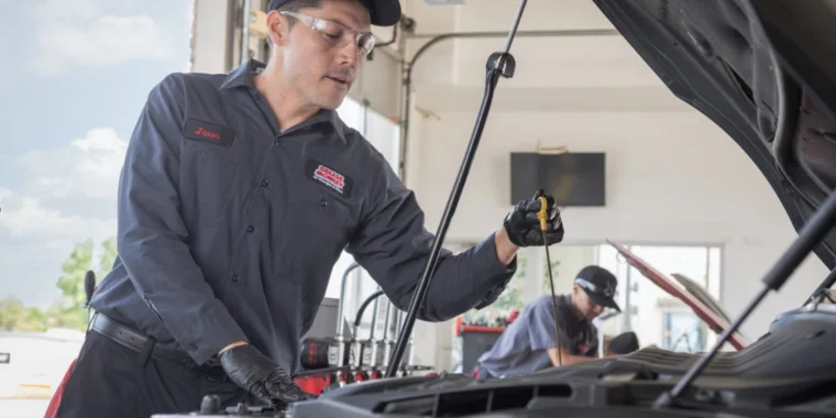 An auto mechanic in uniform and gloves checks the oil level under the hood of a car in a service garage in Idaho Falls, with another worker visible in the background, ensuring engine longevity with every oil change.