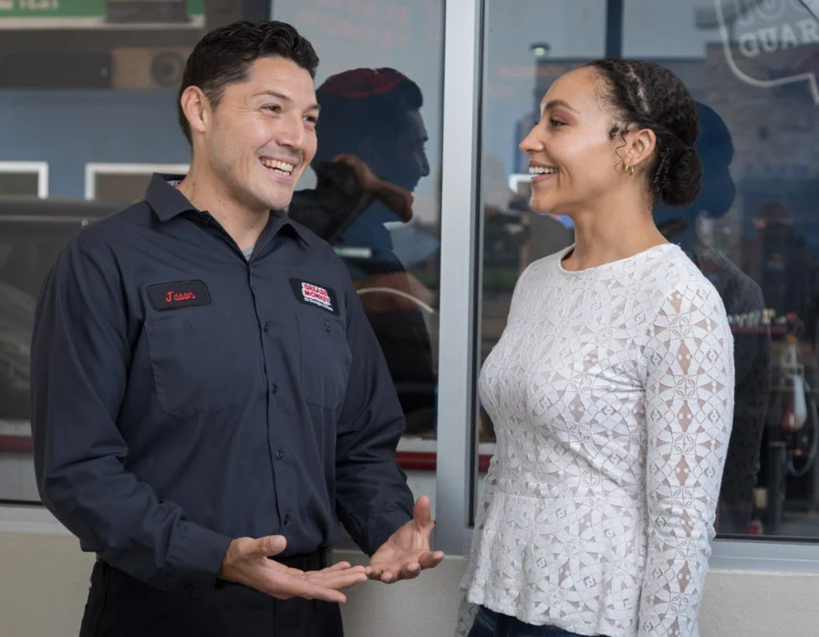 A man in a work uniform chats and gestures while smiling at a woman in a white long-sleeve top; they appear to be having a friendly conversation about vehicle maintenance indoors in Pocatello, Idaho.