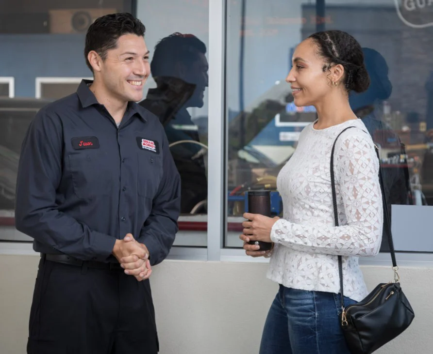 A man in a work uniform smiles and talks with a woman holding a coffee cup outside a building, sharing tips about vehicle safety.