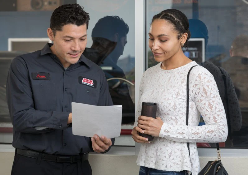 A man in a Grease Monkey work uniform shows a sheet of paper to a woman with a travel mug and backpack by the Grease Monkey, 2100 Channing Way, Idaho Falls window—perhaps discussing a synthetic blend oil change near me to ensure engine longevity.