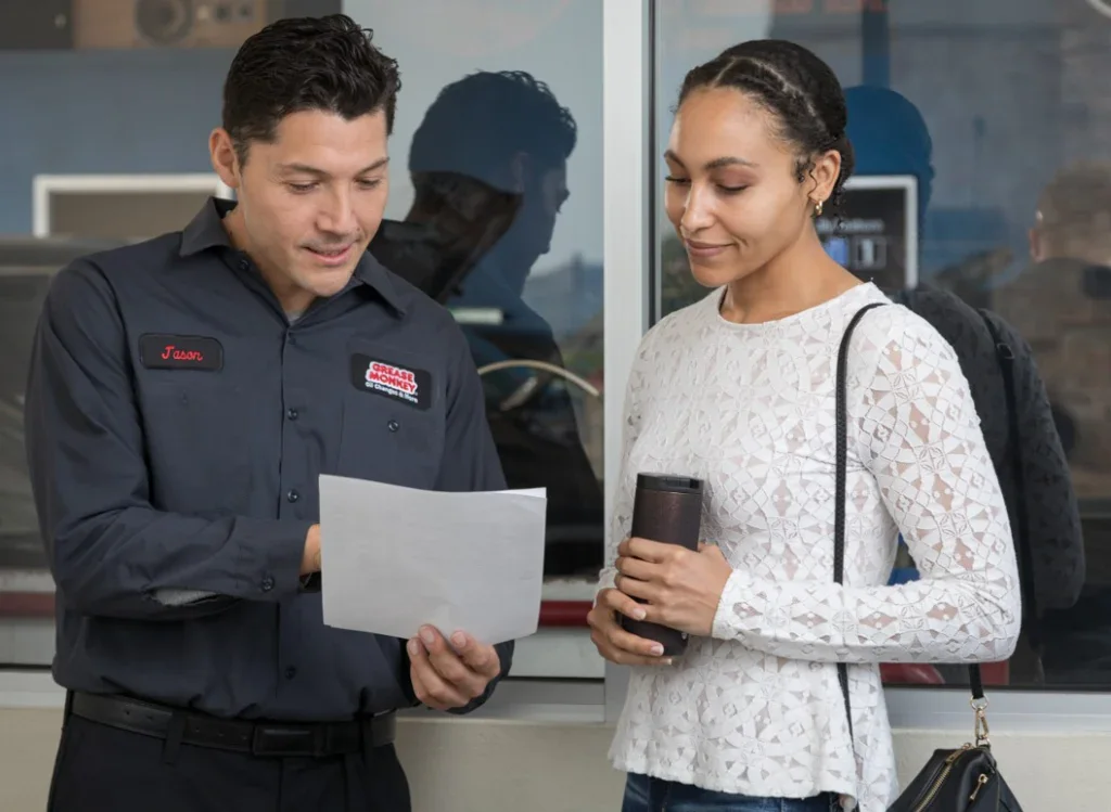 A man in a work uniform shows a document to a woman holding a travel mug near a window, discussing engine protection after her Grease Monkey oil change in Twin Falls, Idaho.