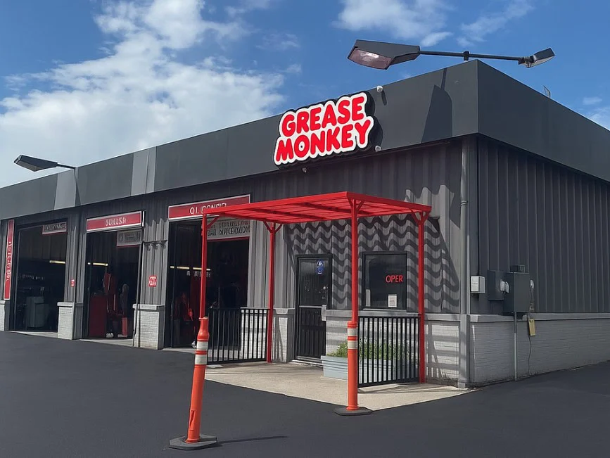 A Grease Monkey auto service shop in Idaho Falls with open service bays, a red awning, and two orange traffic cones in front under a partly cloudy sky, ready to handle your engine maintenance needs.