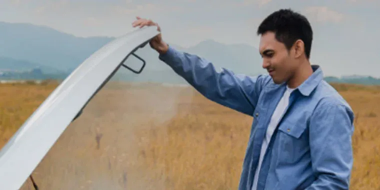 A man standing outdoors in a field near Twin Falls, Idaho, opens the hood of a steaming car, appearing to inspect the engine for potential issues and ensure proper engine protection.