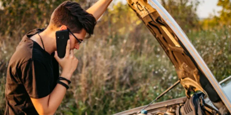 A person stands by a car with the hood open, talking on the phone, possibly seeking help for a vehicle breakdown in an outdoor setting.