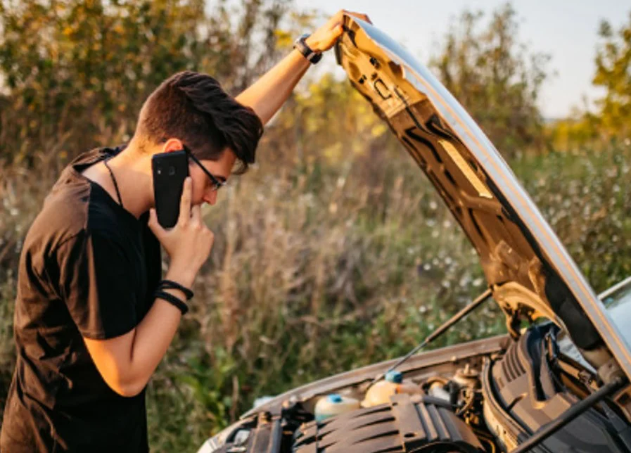 A person stands by a car with the hood open, talking on the phone, possibly seeking help for a vehicle breakdown in an outdoor setting.