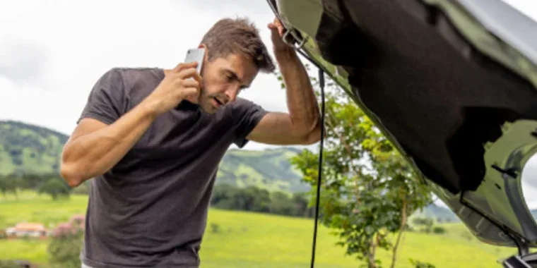 A man stands outdoors looking under the hood of a car while talking on his phone, appearing concerned—he might be searching for a quick oil change in Pocatello.