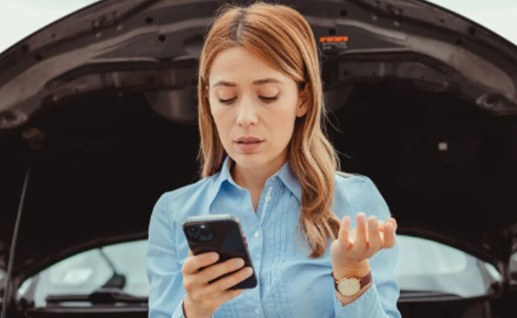 A woman stands in front of a car with its hood open, looking at her phone with a concerned expression—perhaps searching for vehicle maintenance tips to ensure family safety.