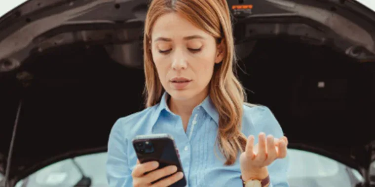 A woman stands in front of a car with its hood open, looking at her phone with a concerned expression—perhaps searching for vehicle maintenance tips to ensure family safety.