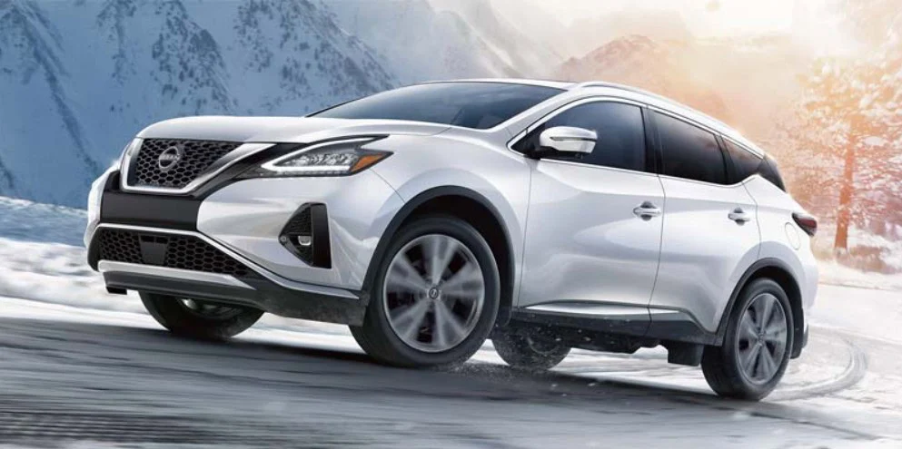 A white SUV is driving on a snowy road with mountains and a cloudy sky in the background, showcasing the importance of engine protection. Trust Grease Monkey oil change in Twin Falls Idaho to keep your vehicle ready for any adventure.